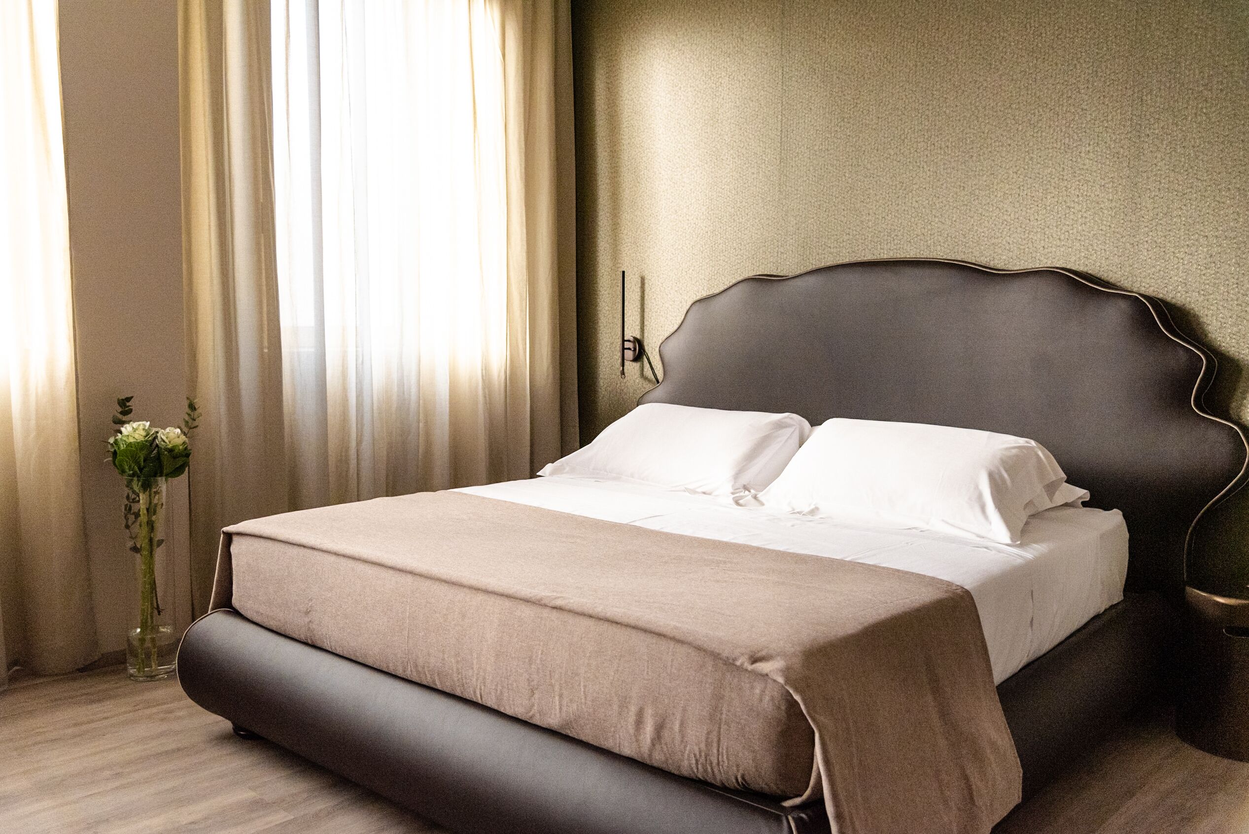 Una cama bien hecha con un cabecero oscuro, un florero con flores y una ventana con cortinas.