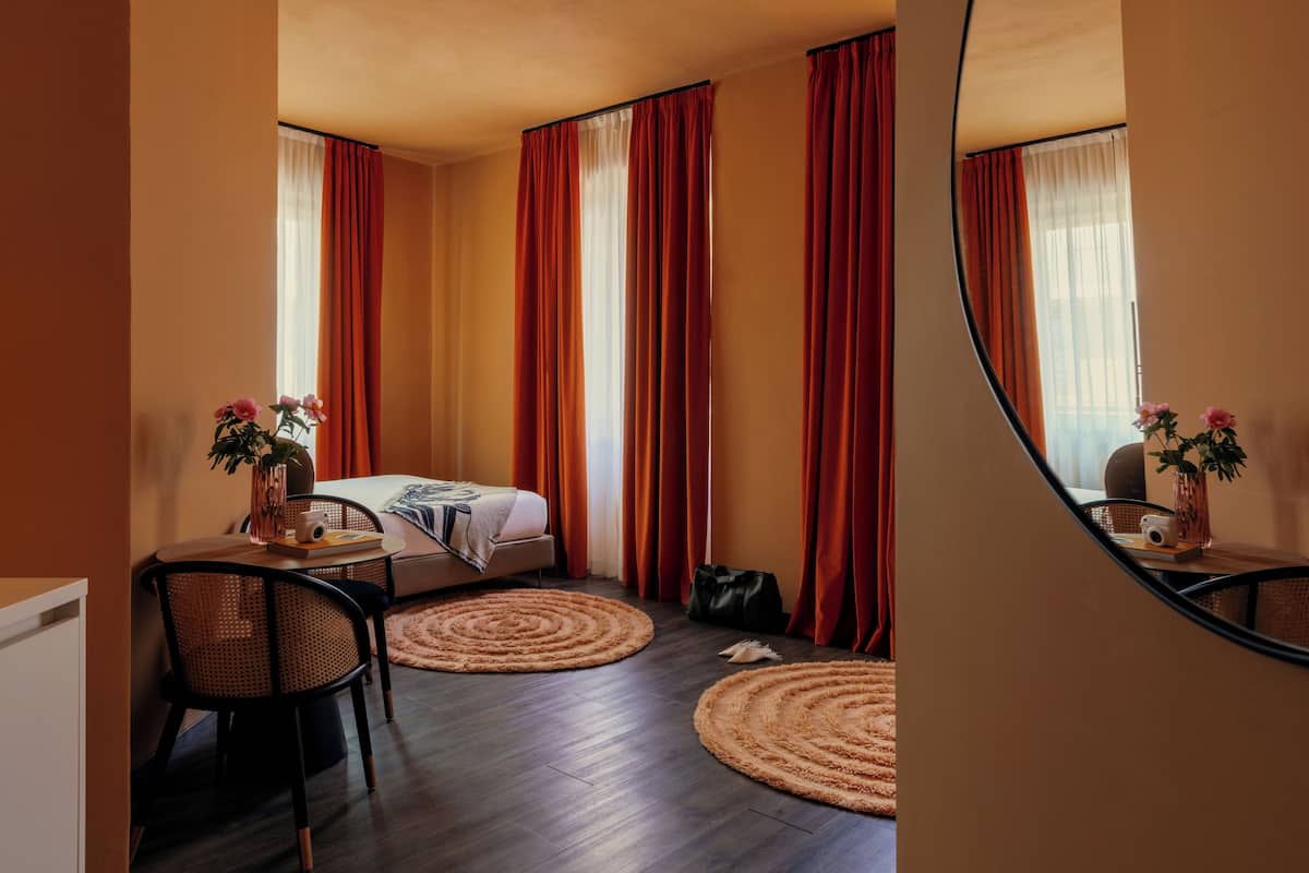 Une chambre d’hôtel avec un lit, un bureau et une chaise, une petite table avec un vase de fleurs et un miroir au mur.