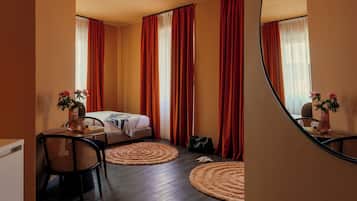A hotel room with a bed, a desk with a chair, a small table with a vase of flowers, and a mirror on the wall.