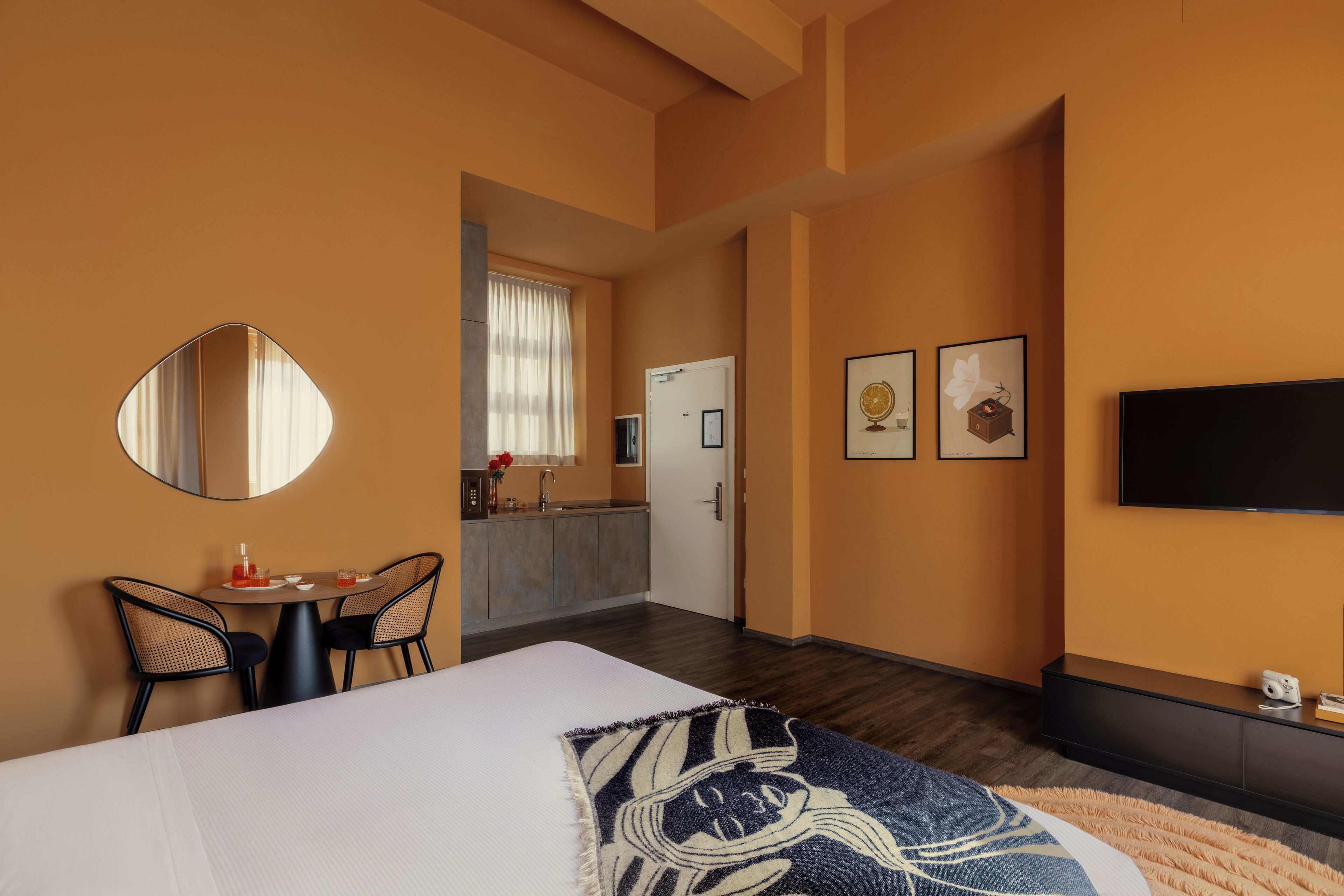 Une chambre d’hôtel comprenant un lit, un coin repas avec des chaises, une cuisine, une télévision et des tableaux encadrés au mur.