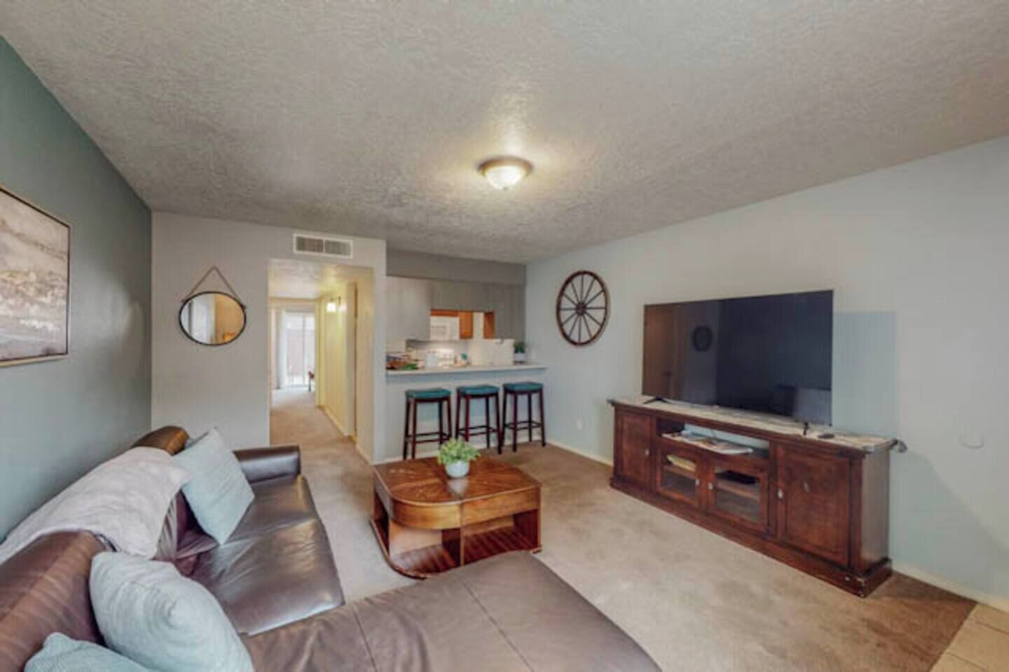 A living room with a sofa, coffee table, TV stand, and kitchen area in the background.