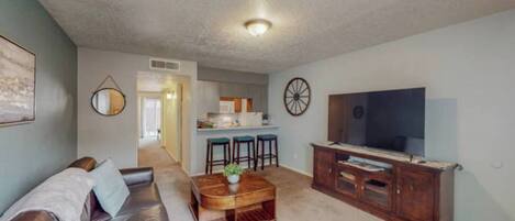 A living room with a sofa, coffee table, TV stand, and kitchen area in the background.