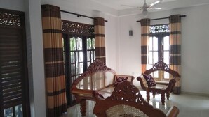 A spacious room with a high ceiling, a ceiling fan, and a large window with curtains. There is a dining table with chairs and a decorative wall panel. - sinhagiri hotel and restaurant (Tangalle)