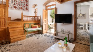 Amplia sala de estar con una escalera de madera, un banco verde, un televisor grande y un área de comedor visible a través de una puerta abierta.