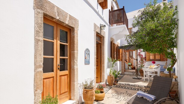 Un patio estrecho con una puerta de madera, plantas en macetas, una alfombra estampada y una silla de lounge.