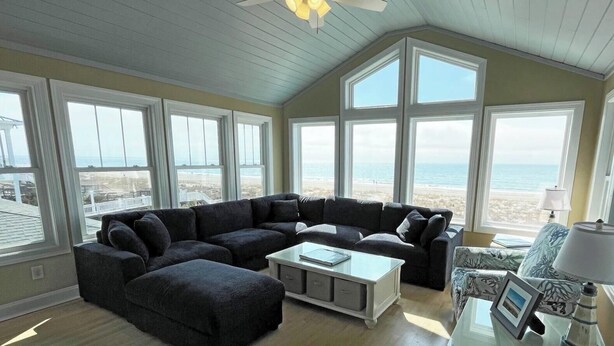 Een kamer met een ruime zithoekbank, een klein tafeltje en een lamp. Er zijn meerdere ramen met uitzicht op het strand.