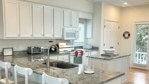 Una cocina con gabinetes blancos, encimera de granito, microondas integrado, estufa, lavabo y una zona de bar con sillas blancas.