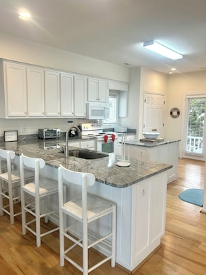 A kitchen with white cabinets, a granite countertop, a built-in microwave, a stove, a sink, and a bar area with white chairs. - 5 bedrooms, 5 bathrooms. Oceanfront with oceanside screened in porch and pet friendly. (Brunswick)