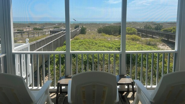 Un balcón con vista a la playa, barandas de madera y algunas sillas.