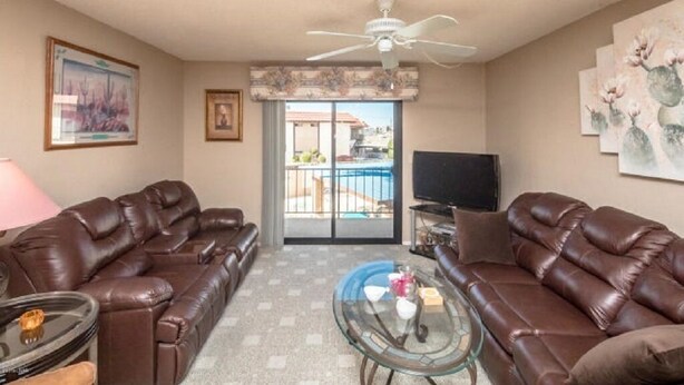 A living room with two brown leather sofas, a glass coffee table, a ceiling fan, a TV, and a door leading to a balcony with a pool view.