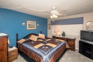 A bedroom with a bed, a ceiling fan, a nightstand, a dresser, a lamp, a TV, and a window with blinds.