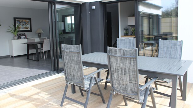 A modern outdoor dining area with a table and chairs, a potted plant, and a view through large sliding glass doors into a kitchen and dining space.