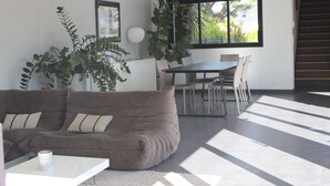 A modern living room with a brown sofa, a dining table with chairs, large windows, and a staircase.