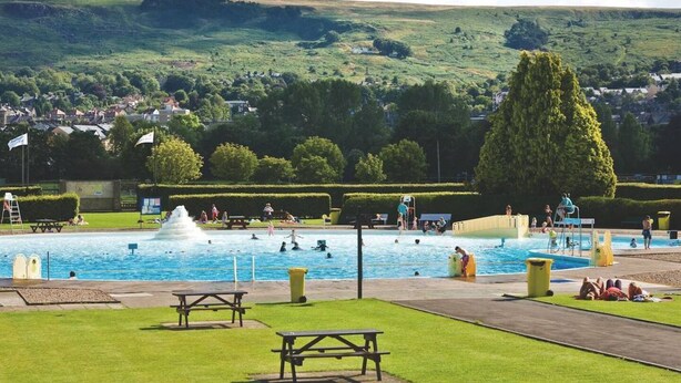 Una piscina con una fontana, circondata da un'area verde con tavoli da picnic e vista su una città sullo sfondo.