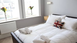 Een netjes opgemaakt bed met witte beddengoed en een kussen met Union Jack, een radiator en een raam met uitzicht op gebouwen.