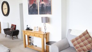 Een woonkamer met een grijze bank, een houten console tafel, een zwarte fauteuil en een rood geruit kussen. Op de muur hangt een klok en boven de console tafel hangen twee ingelijste foto’s.