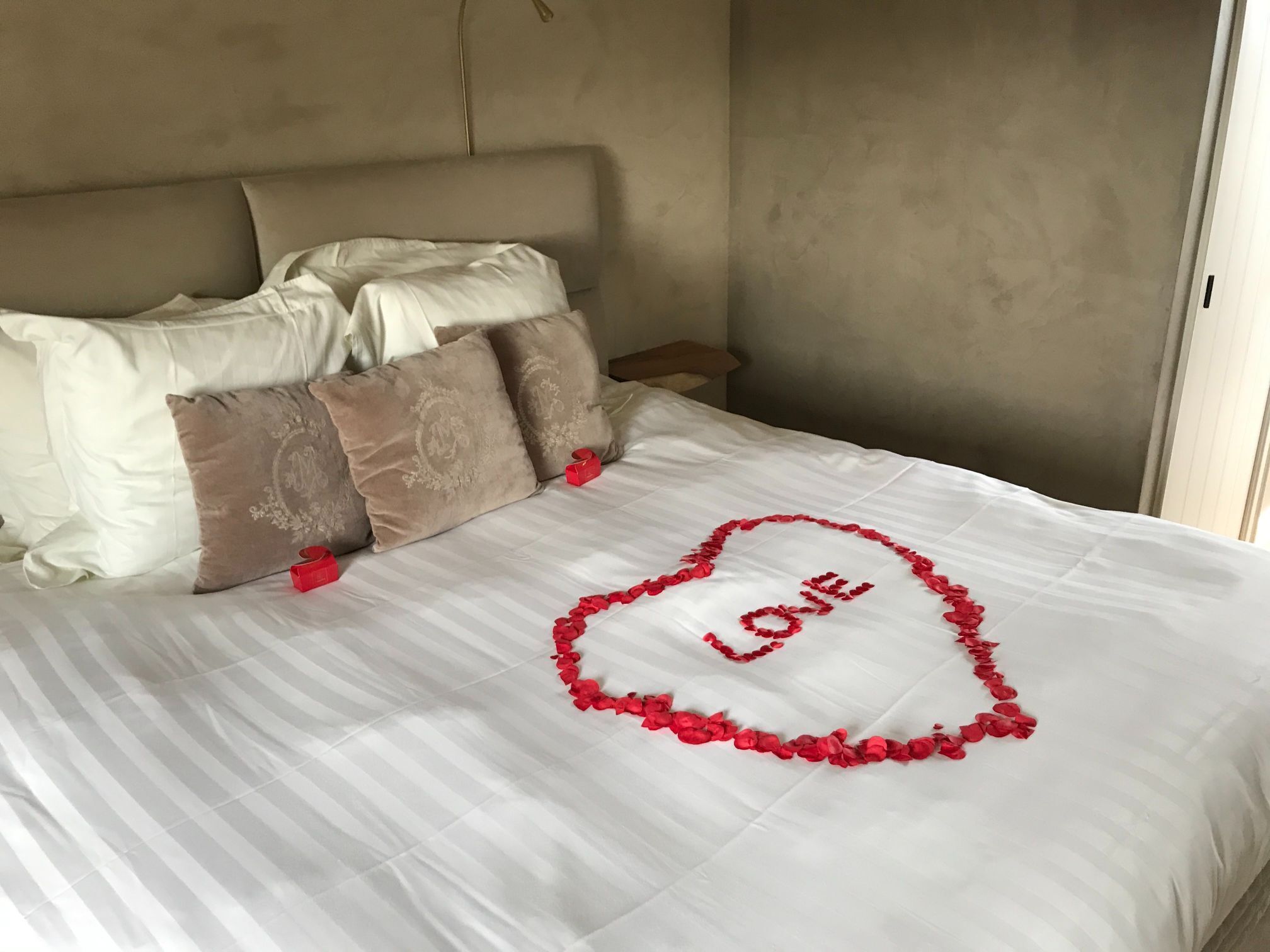 Ein ordentlich bezogenes Bett mit einer herzförmigen Anordnung aus roten Rosenblättern, die das Wort 'LOVE' auf der Bettdecke bilden.