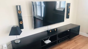 A modern living room with a flat-screen TV mounted on a black entertainment unit, a white floor lamp, and a white speaker on the left. - Canal Waterfront Townhouse (Leeds)