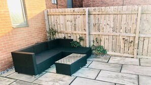 A patio with a dark-colored sectional sofa and a small table, a wooden fence, and a brick wall. - Canal Waterfront Townhouse (Leeds)