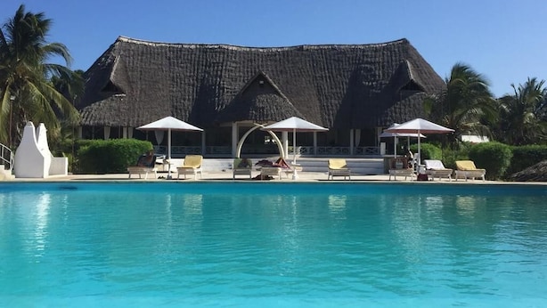 A swimming pool with lounge chairs and umbrellas, a thatched-roof building, and palm trees.