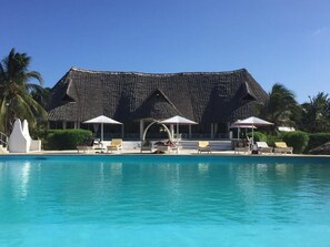 Una piscina con sillones reclinables y sombrillas, un edificio con techo de paja y palmeras.