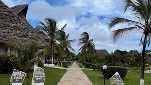 A stone pathway leading to a resort area with palm trees and a thatched-roof building. Directional signs point to 'Disco Pizzeria Bocce' and 'Chui Restaurant'.