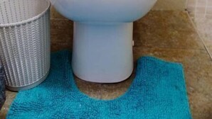 Uma sanita branca numa casa de banho com azulejos bege, um tapete de casa de banho azul e uma cesta de metal.