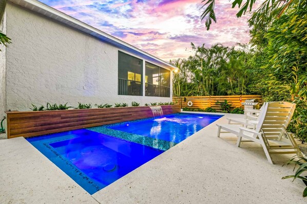 A modern outdoor swimming pool with a wooden deck and lounge chairs, surrounded by lush greenery and a white building with large windows. - Family Vacation Home with Pool and Spa, 2-min walk to Beach - Stone's Throw (Lido Key)