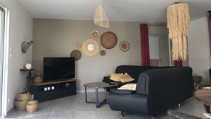 A modern living room with a black sofa, a small round table, and a TV on a wooden stand. There are woven baskets on the floor and a variety of woven wall decorations. - Holiday villa with pool in Vallon Pont d'Arc, near the Ardèche river (Vallon-Pont-d'Arc)