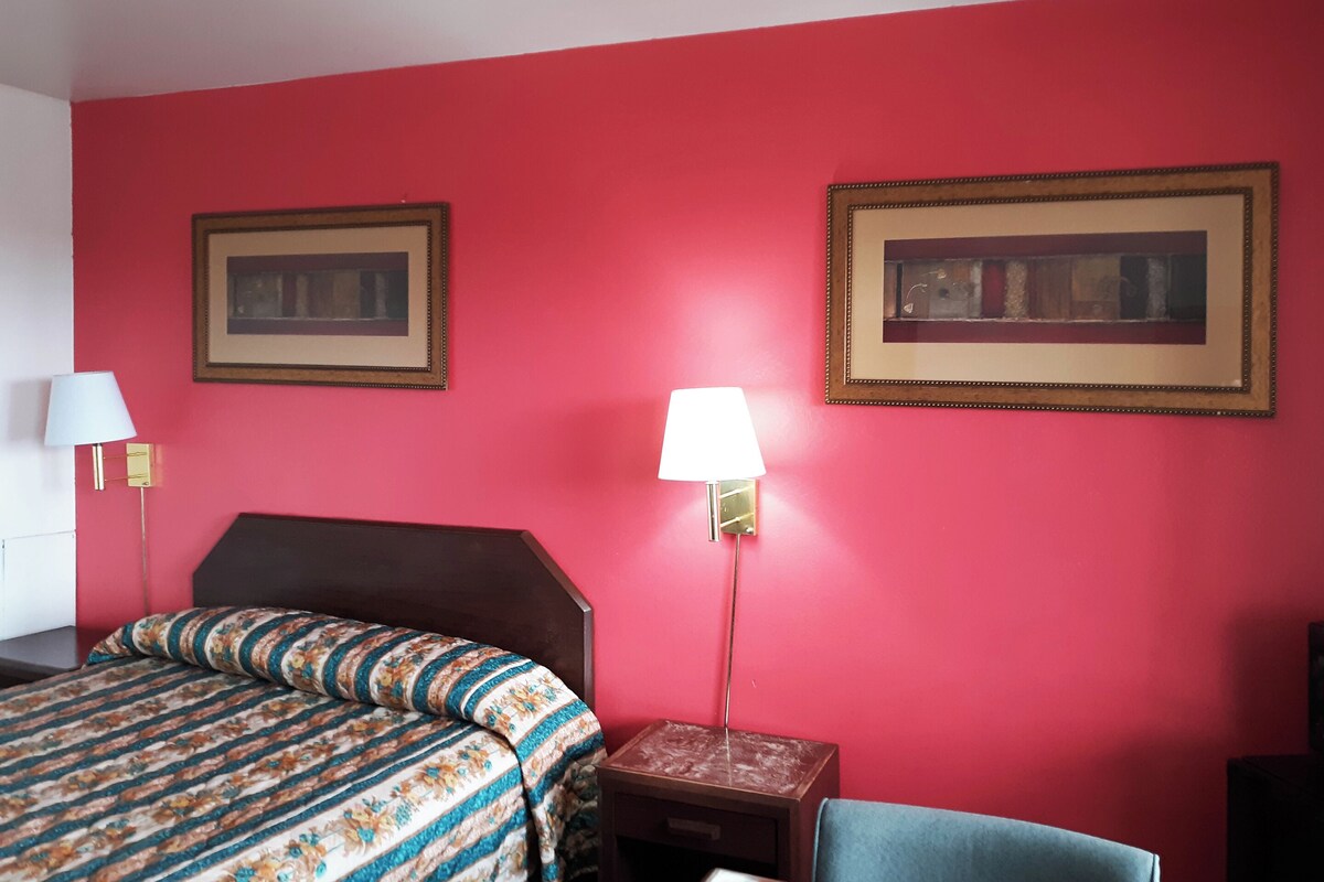 A hotel room with a red wall, a bed with a floral bedspread, two framed pictures, a bedside lamp, and a chair.