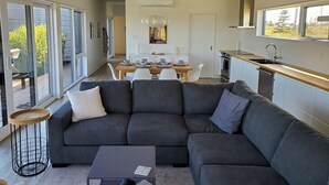 En moderne stue med en hjørnesofa, et spiseområde med et bord dækket til fire personer, og et køkken med vask og komfur.