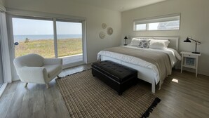 Et soveværelse med en stor skydedør til en balkon, en seng med hvide sengetøj, en sort fodskammel og et natbord med en lampe.