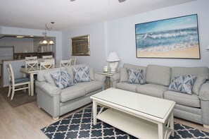 Una sala de estar con un sofá gris, cojines a juego, una mesa de centro blanca y una alfombra estampada. En la pared, un cuadro con una escena de playa.
