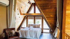Un dormitorio con techo inclinado, una cama con ropa de cama blanca, un sillón de cuero marrón y ventanas grandes.
