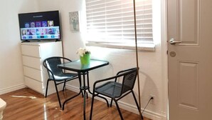 Een kleine kamer met houten vloer, een witte deur, een raam met jaloezieën, een tafel met twee stoelen, een tv op een standaard en een staande lamp.
