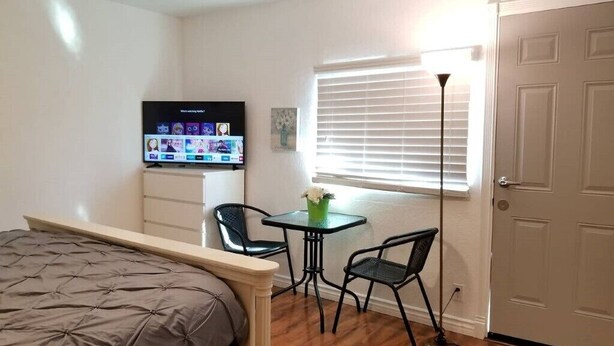 Een slaapkamer met een bed, een aan de muur bevestigde televisie, een klein tafeltje met twee stoelen, een raam met zonwering en een deur.