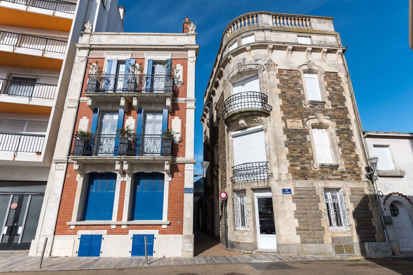 Zwei Gebäude mit unterschiedlichen Baustilen, eines mit Balkon und blauen Fensterläden, das andere mit einem Eckturm und vergitterten Fenstern.