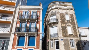 Deux bâtiments aux styles architecturaux différents : l’un possède un balcon et des volets bleus, l’autre, une tourelle d’angle et des fenêtres grillagées.