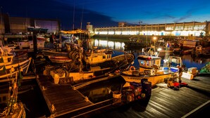 Un port de plaisance de nuit, avec des bateaux amarrés, un grand bâtiment en arrière-plan et une jetée en bois.
