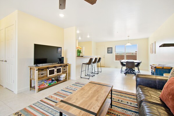 Een woonkamer met een televisie, een houten entertainmentkast en een salontafel. Er is een eethoek met een tafel en stoelen, en een keuken op de achtergrond.
