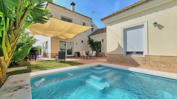 Une piscine dotée d’un parasol, une terrasse avec une table et des chaises, et un bâtiment avec un toit en tuiles.