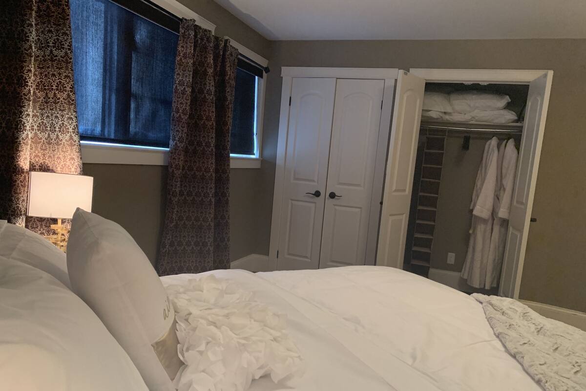 A bedroom with a bed, a window with curtains, a wardrobe, and a closet with hanging clothes.
