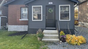 A small, two-story house with a dark gray exterior, a black front door, and a number "301" sign above the door. There are two windows on the front facade, a small porch with steps, and a variety of potted plants in front of the house. - Legacy Suites (Sudbury)