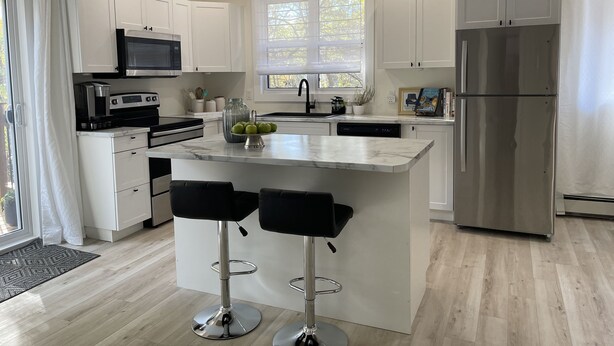 A modern kitchen with white cabinets, a stainless steel refrigerator, a central island with bar stools, and pendant lighting. - Legacy Suites (Sudbury)