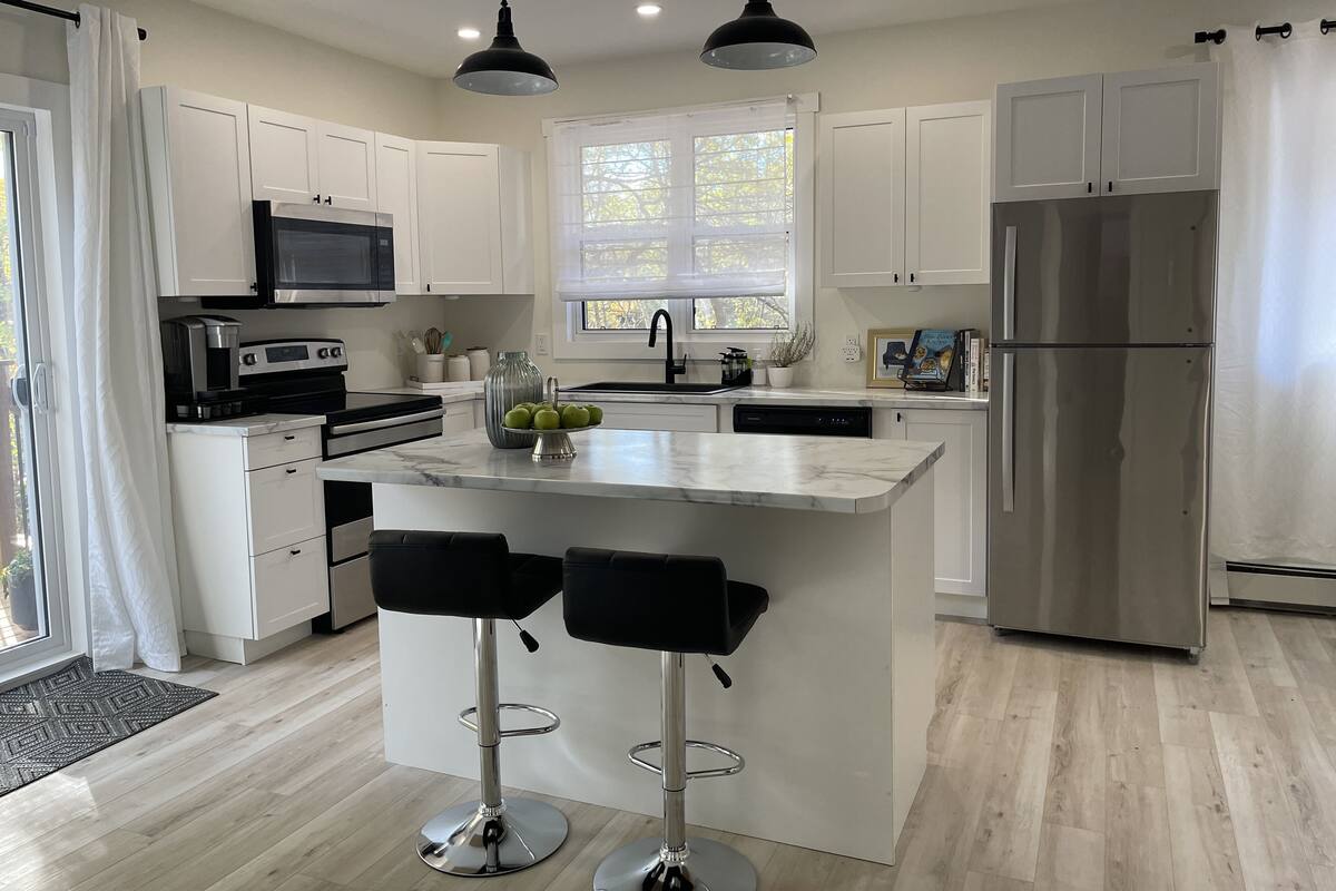 A modern kitchen with white cabinets, a stainless steel refrigerator, a central island with bar stools, and pendant lighting.