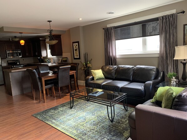A modern living room with a leather sofa, a glass coffee table, and a dining area with a kitchen in the background.