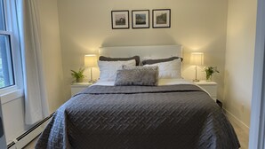A neatly made bed with a grey quilt, two bedside lamps, and a window with white curtains. - Legacy Suites (Sudbury)