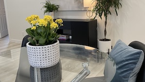 A modern dining area with a glass table, black chairs, a blue cushion, a potted plant, and a wall-mounted painting. - Legacy Suites (Sudbury)