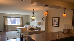 A modern living room with a glass dining table, beige sofas, and a large kitchen island. - Legacy Suites (Sudbury)