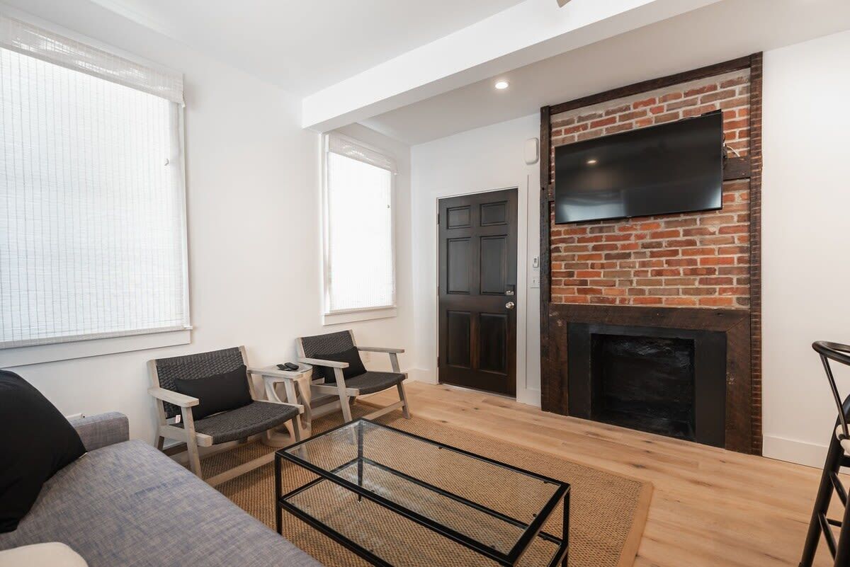 A modern living room with a brick fireplace, a flat-screen TV mounted on the wall, and a seating area with a sofa and two chairs.